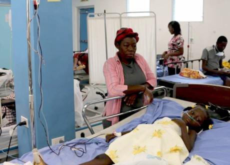 A young Haitian girl wears a nasal cannula while lying on a hospital bed in the emergency room. Her mother leans over her bed.