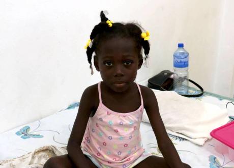 A young Haitian girl wearing a pink tank top sits cross-legged on a hospital bed.