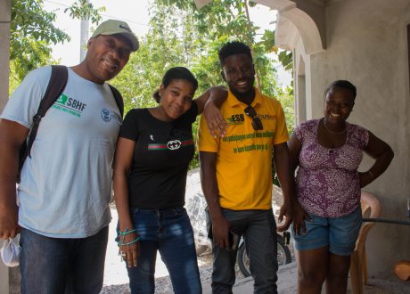 A Haitian woman stands outside of her home with the clinians and physical therapists who helped her learn to walk again. All four people smile at the camera.