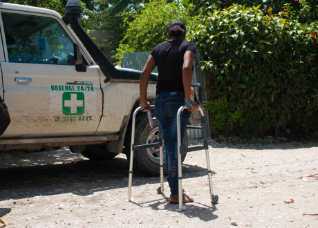 A Haitian woman stands with a walker on a dusty road. Her back is to the camera and a white ambulance stands to her left.