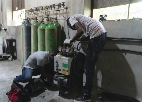 Two Haitian biomedical repair technicians install an oxygen concetrator. The machine is a large dark box placed against a cement wall next to tall green oxygen bottles.