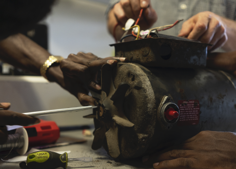 A close-up of a motor for an oxygen concentrator. Two mens' hands are placed on the motor, in the process of fixing it.