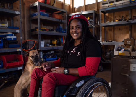 A Haitian woman in a wheelchair sits in the middle of an equipment workshop. A light-colored dog is peeking out from over her leg. The woman smiles.
