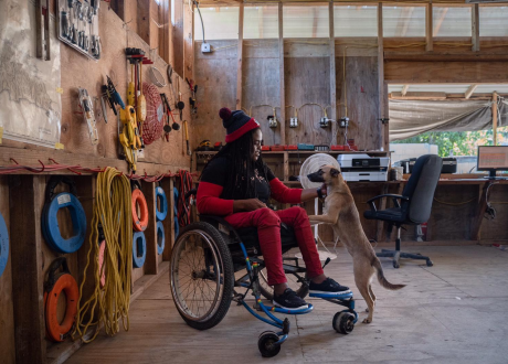 A Haitian woman in a red wheelchair sits in the middle of an equipment workshop. A light-colored dog jumps up to lean on her legs.