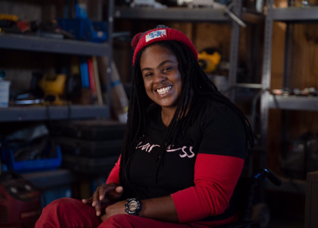 A Haitian woman sits in a wheelchair in an equipment workshop. She smiles brightly at the camera.