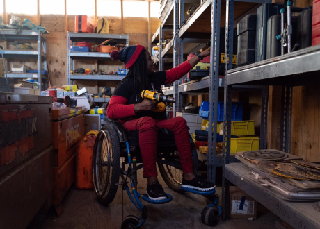A Haitian woman in a wheelchair is in an equipment workshop. She reaches into a metal shelving unit for a tool.