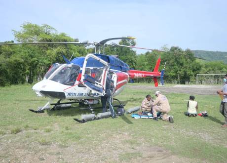 A helicopter lands in a grassy field. Two medics in tan jumpsuits tend to a patient on the ground outside of the helicopter.