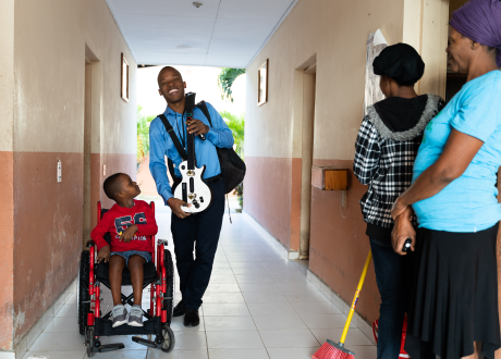 A young Haitian boy in a wheelchair looks up at a hospital worker, who stands and smiles to his right. Two smiling women look on.