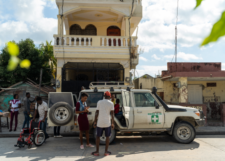 A white ambulance stands in front of a tall, narrow apartment complex in Les Cayes, Haiti. A crowd of people is gathered around the vehicle.