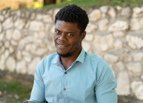 A Haitian man sits in front of a low stone wall. He wears a light blue button down shirt and has short, dark hair. He smiles directly at the camera.