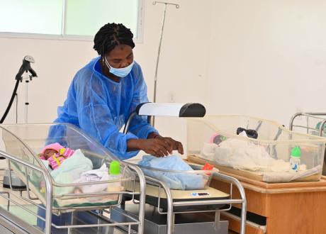 In a hospital, a Haitian woman in a blue gown and surgical mask arranges a blanket on a newborn situated in a bassinet. Two additional newborns rest, one on each side of her.