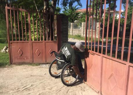 A Haitian man in a wheelchair tends to a reddish-pink metal gate outside on a dusty road.