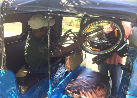 A Haitian man sits in a taxi and pulls his wheelchair into the cab with the help of a driver.
