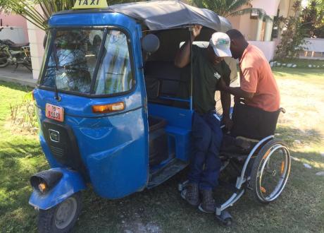 A Haitian man in a wheelchair climbs into a blue three-wheeled taxi with the help of a driver.
