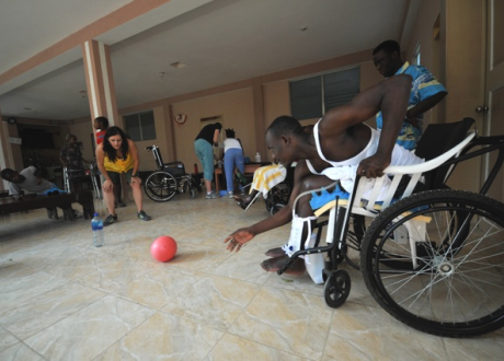 A Haitian man in a wheelchair bends at the waist to retrieve a red ball that is rolling towards him