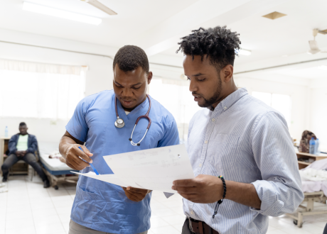 A Haitian clinician in light blue scrubs reviews a series of paper with a colleague, who stands to his right.