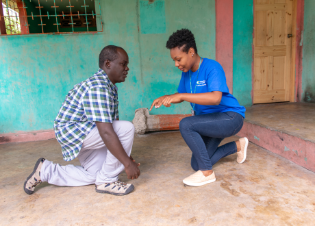 A female Haitian physical therapist crouches to provide physical therapist to a male patient at his home; the man is also crouching.