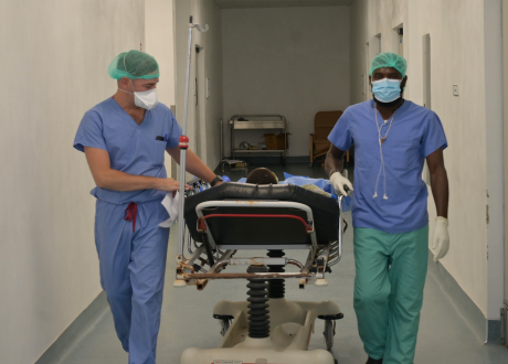 2 surgeons bring a patient on a gurney down a hall to the OR. They wear blue scrubs, medical masks, and hair nets.