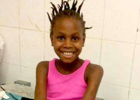 A young Haitian girl smiles brightly at the camera while seated on a hospital bed in front of a tile wall.