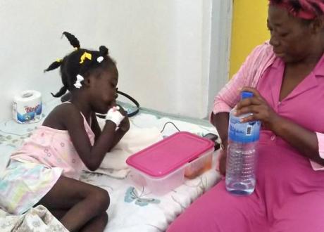 A young Haitian girl sits on a hospital bed. Her mother sits next to her in a chair.