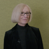 A white woman stands in front of a light green wall. She is shown from the waist up. She wears a black velvet jacket and black turtleneck. She has black glasses and a short blonde bob haircut.
