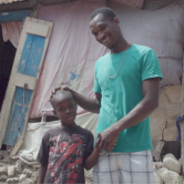 A young Haitian boy stands next to his dad outside. They stand in front of a home destroyed in the 2021 earthquake. The little boy has short hair and wears a spider man t-shirt. His father holds his hand and touches his head gently. His father wears a teal t-shirt and white and black striped shorts. They are smiling.