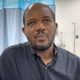 A Haitian doctor stands in front of a curtain in the surgica center. He wears a navy blue button-down shirt that has small white polka dots on it. He has short hair and a short beard.