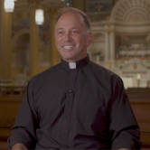 A priest sits in a pew in a large church. He wears a black shirt and a white clerical collar. He has short hair. He smiles at the camera. He is shown from the waist up.