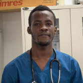 A man Haitian clinician stands in front of a cabinet. He wears a dark blue scrubs top and has a stethoscope around his neck. He has short dark hair.