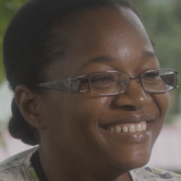 A Haitian woman nurse looks to the right and smiles. She is shown from the neck up. Her hair ist ied back in a bun. She wears rectangular wire-rimmed glasses.