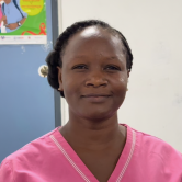 A Haitian woman nurse stands in front of a blue and white wall. She is shown from the shoulders up. She wears a bright pink scrubs top. She looks at the camera and smiles. Her braided hair is tied back.