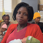 A patient named Nana sits in the community health room surrounded by other mothers and their babies. Nana has short braided hair. She wears a red top and cardigan and cradles her baby, who is wrapped in a blanket.