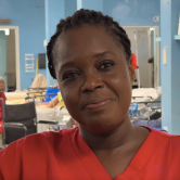 A Haitian Nurse is pictured in the emergency room from the shoulders up. She is smiling. Her hair is braided and tied back and she is wearing red scrubs.