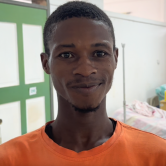 A Haitian man stands in a hospital room. He is shown from the shoulders up. He has very short hair and wears an orange T-shirt. He is smiling.