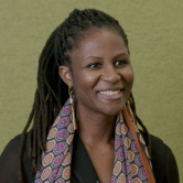 A dark-skinned woman stands in front of a green background. Her long braided hair is tied back. She wears a black shirt and a brightly patterned scarf. She smiles at the camera.