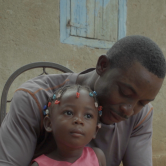A Haitian man sits in a chair outside with his daughter in his lap. The man has short hair and wears a trey t-shirt. The little girl wears a pink dress and has braided hair with barrettes at the ends.