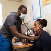 A Haitian doctor stands checks a female patient's breathing with a stethescope on her chest. The patient sits in a chair next to a desk.