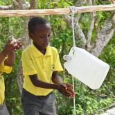 A young boy and girl in yellow school polo shirts wash their hands using a tippy tap device.
