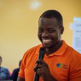 Dr. Galliote from the Kore Sante team stands in a yellow room. He is wearing a bright orange polo shirt with the Kore Sante logo on it. He holds a microphone and smiles brightly.