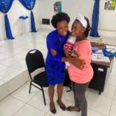 2 Haitian women embrace, smiling, in a white tiled room decorated in blue. One wears a blue dress and the other wears pink and holds an infant.