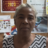 A Haitian woman stands in front of a bulletin board that has several colorful postings on it. Her braided grey hair is tied back. She wears a black and white zig-zag patterned sleeveless top. She is shown from the shoulders up. She smiles at the camera.