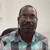 A Haitian man sits in a chair and looks at the camera. He is shown from the shoulders up. He wears glasses with a thin black glasses strap. He has short dark hair and wears a checkered shirt that is black, grey, white, and light blue.