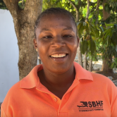 A Haitian woman smiles brightly at the camera. She is standing in front of a tree outside. She wears a bright orange polo shirt with the saint boniface haiti foundation logo embroidered on the left side. She has dark hair that is tied back.