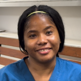 A Haitian woman smiles brightly at the camera. She is sitting on a bench inside. She has dark hair that is loosely tied back, with a thin yellow headband above her forehead. She wears a blue scrubs top.