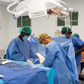 Three Haitian doctors perform a surgical procedure. They crowd around a surgical table wearing light blue scrubs, medical masks, and colorful caps.
