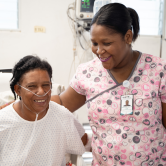 A female Haitian doctor stands next to her patient in the ER. The Doctor puts her arm arount he patient and smiles. The patient wears a white hospital gown and a nasal cannula. The patient also smiles.