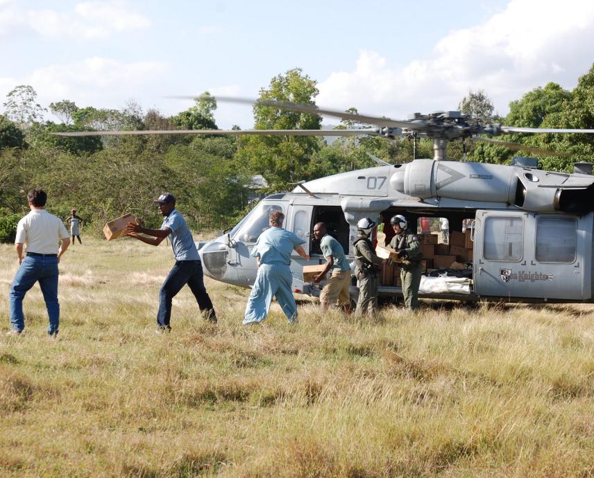 A helicopter sits in a wide field with trees in the background. Doctors and staff memebrs of a hospital unload supplies from the helicopter in an assembly line. 