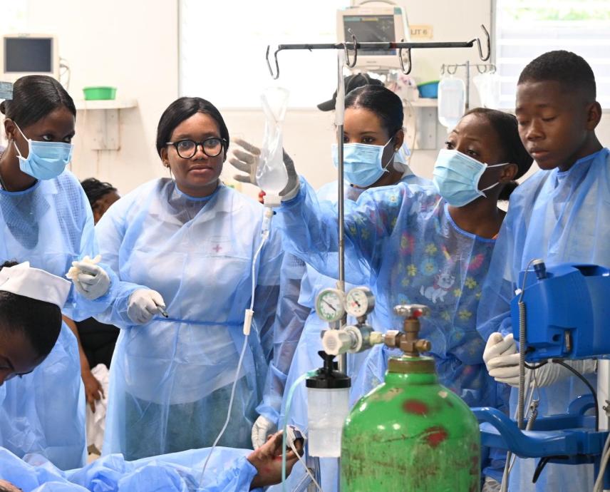 A half-dozen Haitian clinicians in blue medical gowns surround the bed of an ER patient who is receiving IV fluids.