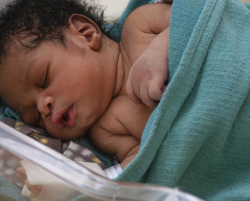 A Haitian baby rests in a bassinet in HEI/SBH's NICU. The baby has dark curly hair and is cuddled into a light blue blanket.