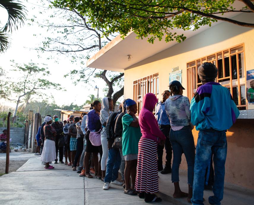 In early-morning light, a line of Haitian men and women stretches from a pale yellow building out through open hospital gates.